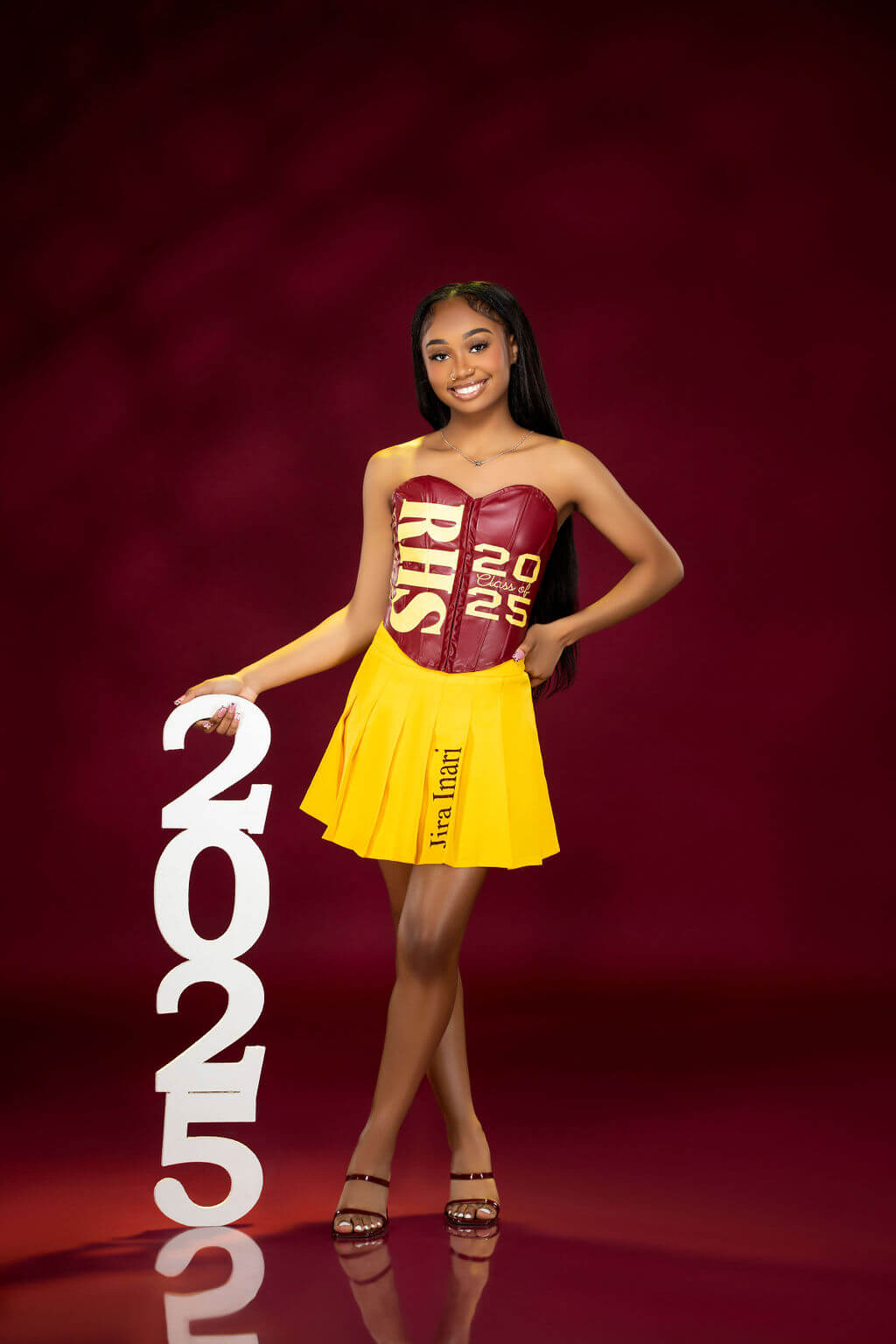 Custom Graduation Outfit with '2025' Prop – A graduate poses in a unique maroon and yellow outfit featuring her school initials and class year while holding a large 