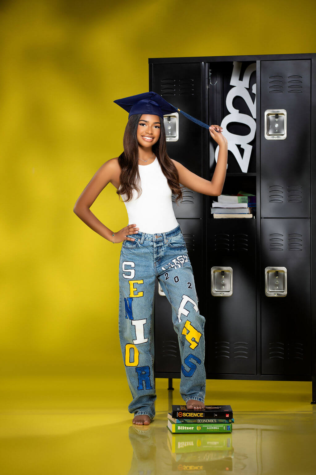 Casual Graduation Look with Lockers – A smiling senior in custom-painted jeans spelling "SENIOR" stands in front of black lockers, holding her graduation cap tassel while the "2025" number from Collageandwood.com is displayed inside a locker.