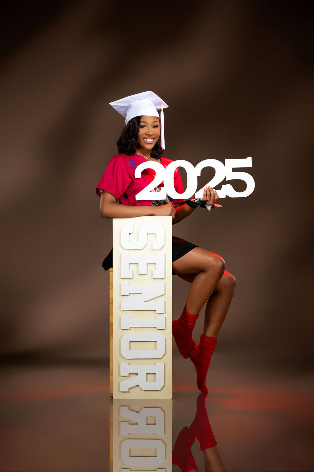 Senior Night Photo with '2025' Prop – A young woman in a red school tee and white cap sits on a tall "SENIOR" prop while holding a "2025" cutout, both props from CollageandWood.com, smiling against a neutral-toned background.