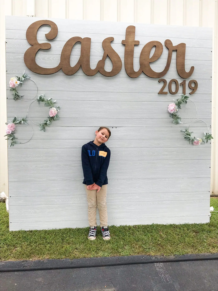 Wooden “Easter” sign hanging on a white backdrop with greenery and flowers, perfect for a church photo wall to celebrate Jesus. Ideal for window displays or Easter shop decor. Add to cart now before it becomes unavailable — checkout with ease and choose the size you prefer. Available at collageandwood.com.